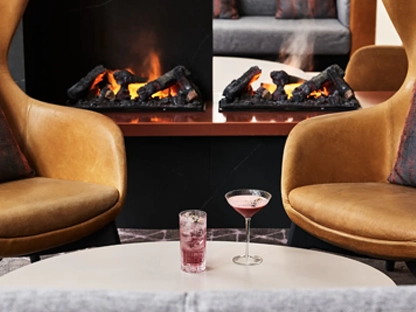 Two leather chairs in front of a fire with a table with two cocktails on it