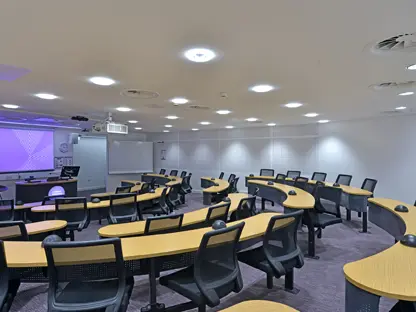 A classroom with long curved desks and chairs surrounding them, facing a presentation screen