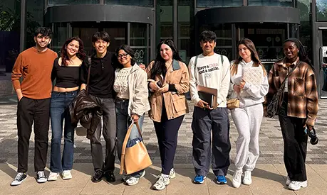 Group of eight young adults standing side by side outdoors in front of the Alliance Manchester Business School building, smiling and casually dressed, some holding notebooks or bags.