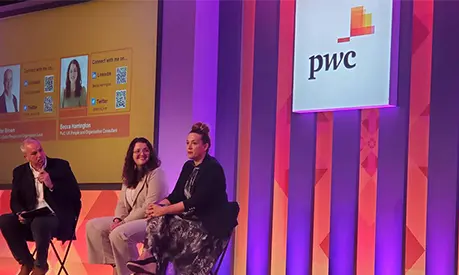 Panel discussion at a PwC event with three speakers seated on stage, PwC logo displayed on a large screen behind them.