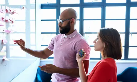 A man and a woman gathered around a whiteboard holding post-it notes and discussing ideas