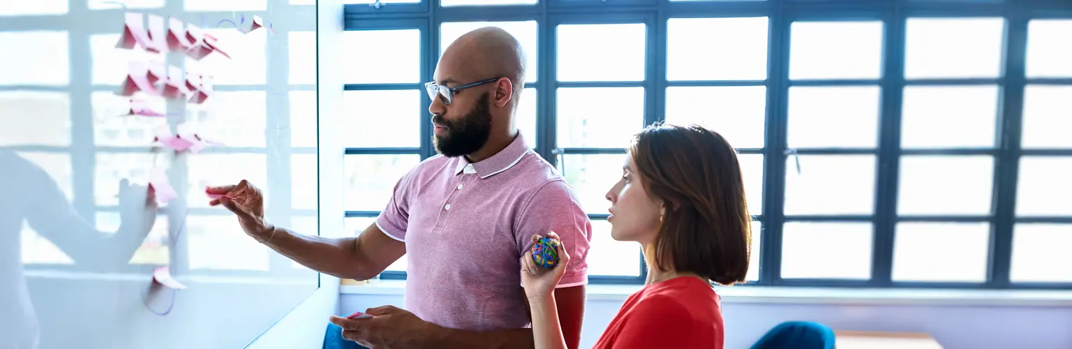 A man and woman gathered around a whiteboard holding post-it notes and discussing ideas