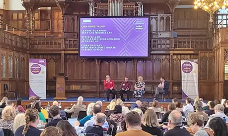 Large audience seated attending the National Conference on Societal Resilience. Four speakers sit on stage beneath a screen while chandeliers hang overhead and natural light streams through tall stained-glass windows.