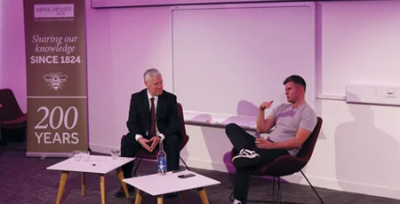 Tom Beahon and Ian King sat in front of some small tables having a conversation at the front of a conference room