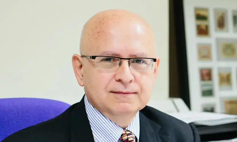 A man in a suit sitting on a purple chair with framed pictures on the wall behind.
