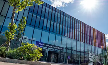 Exterior view of Alliance Manchester Business School building with glass façade and colorful vertical panels, surrounded by greenery on a sunny day.
