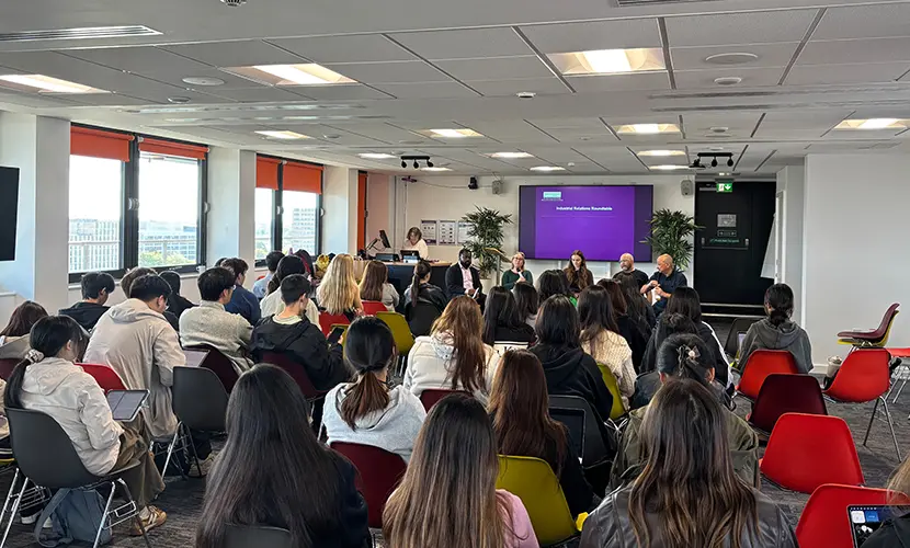 An event with rows of chairs full of students and a panel of five people speaking at the front of the room.