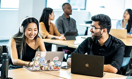 Two students working together on laptops in a seminar room.