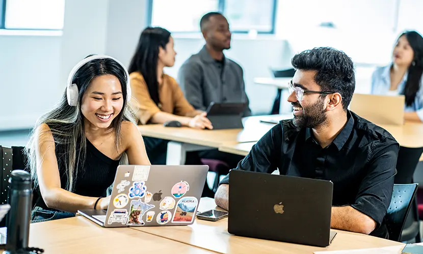 Two students working together on laptops in a seminar room.