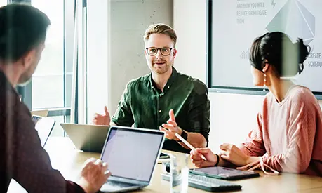 A group of three professionals sat around a table with laptops discussing a group project