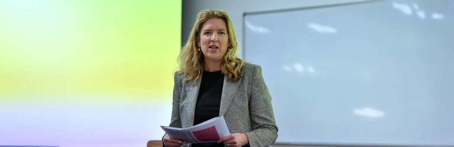 A woman lecturer stands at the front of a classroom holding papers