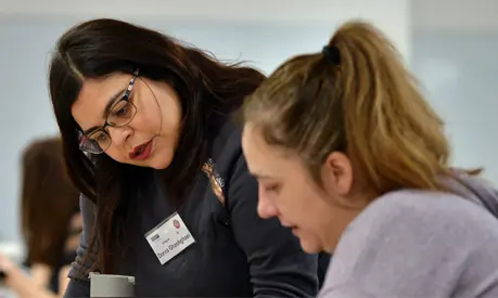 A woman helping another woman with some work in a classroom