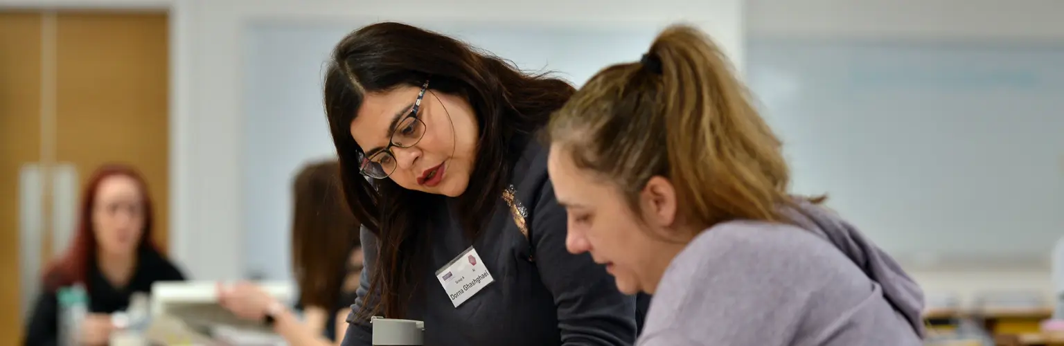 A woman helping another woman with some work in a classroom