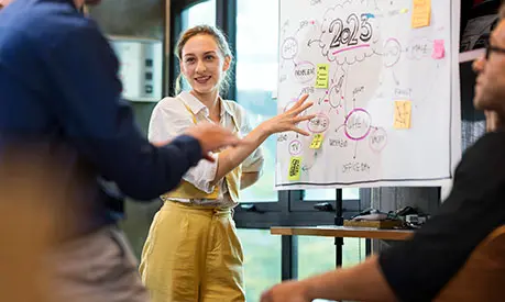 A professionally dressed female standing next to a white board pointing to written ideas in front of her peers